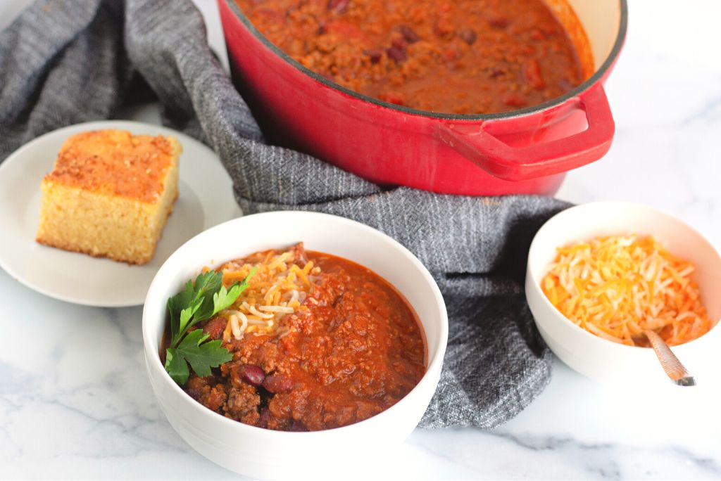 Hearty beef chili bowl with cornbread, cheese, and red dutch oven